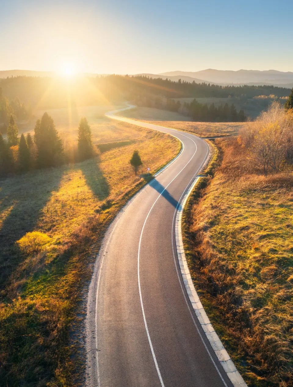 An aerial view of open stretch of road in the countryside, with the sun shining in the background.