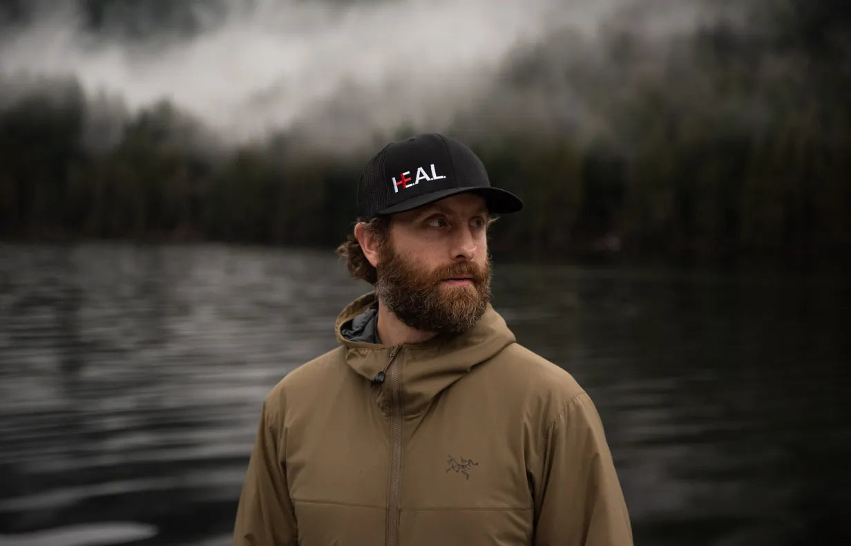 A portrait of Jordan Birch standing in front of a lake, wearing a HEAL baseball cap.